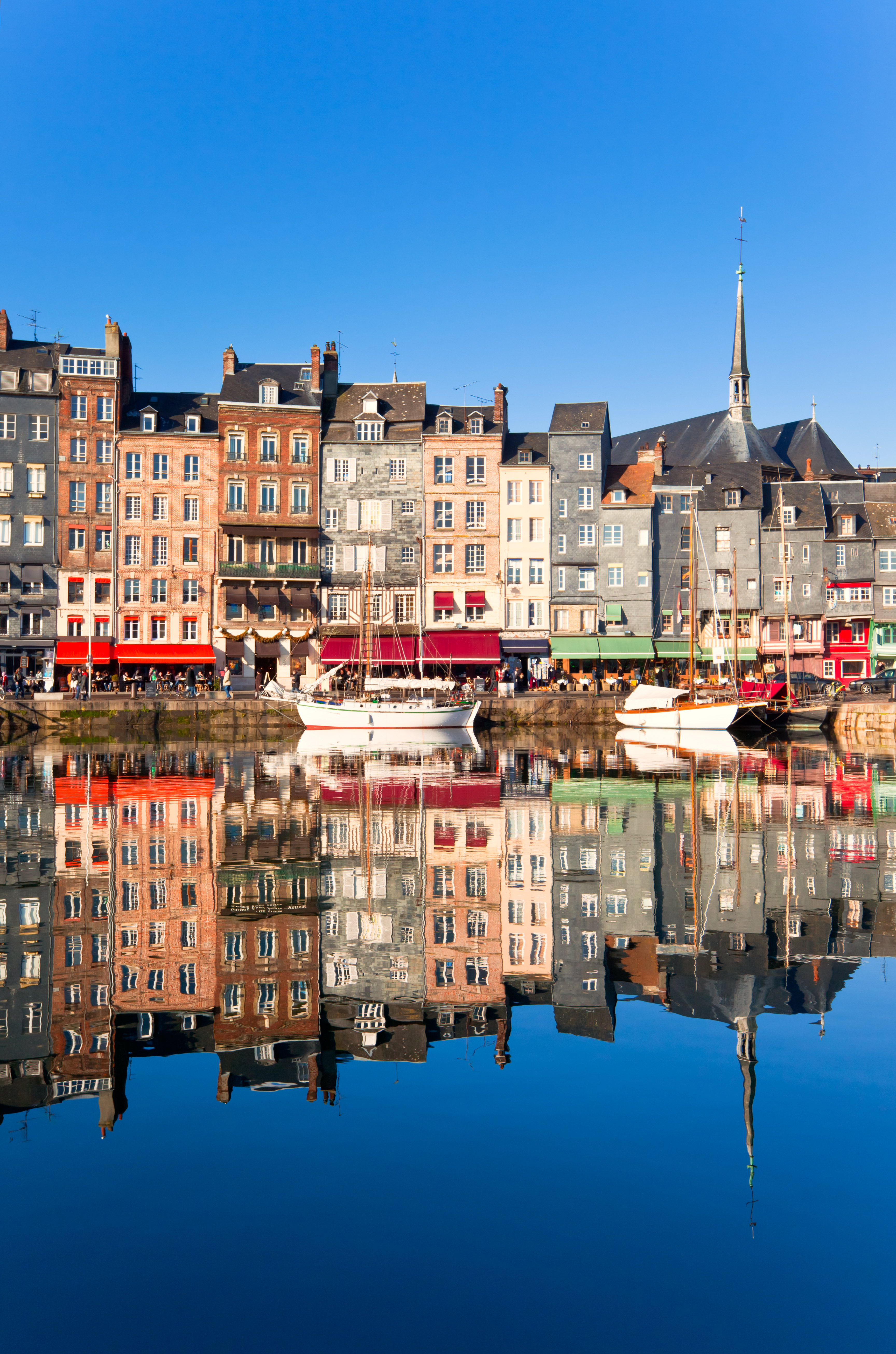 Port of Honfleur with colorful historic buildings and sailboats in the foreground, under a clear blue sky.