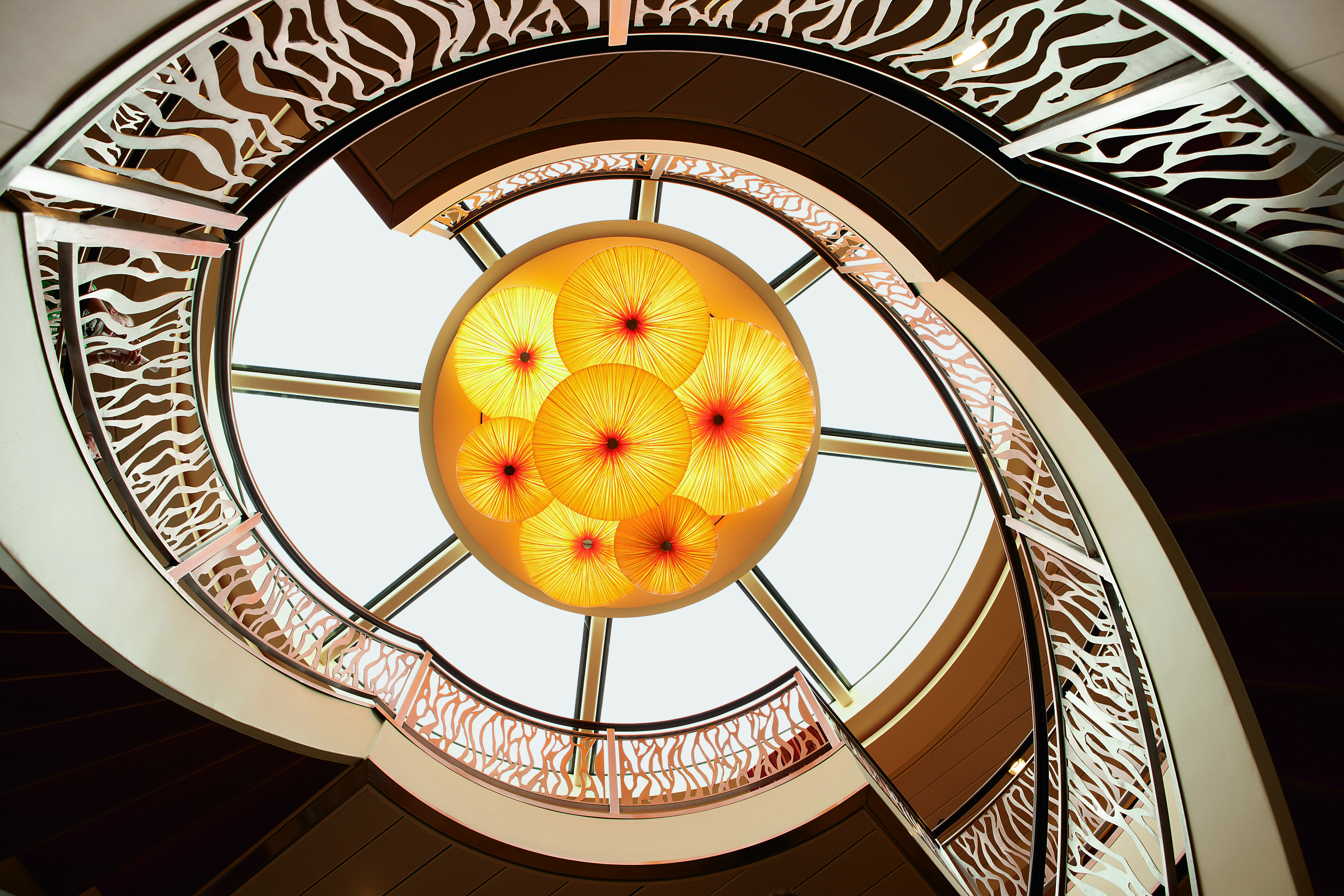 Looking up at a spiral staircase with a decorative railing and a large skylight with yellow, flower-shaped lamps in the center.